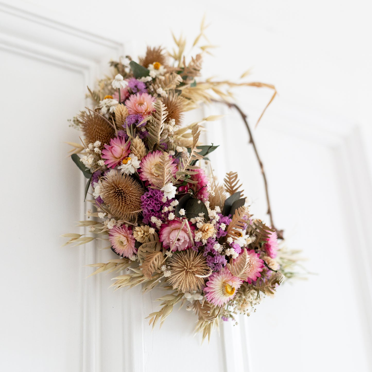 side view of a pink , green and golden brown wreath. hung on a white door. wreath is half covered in dried flowers