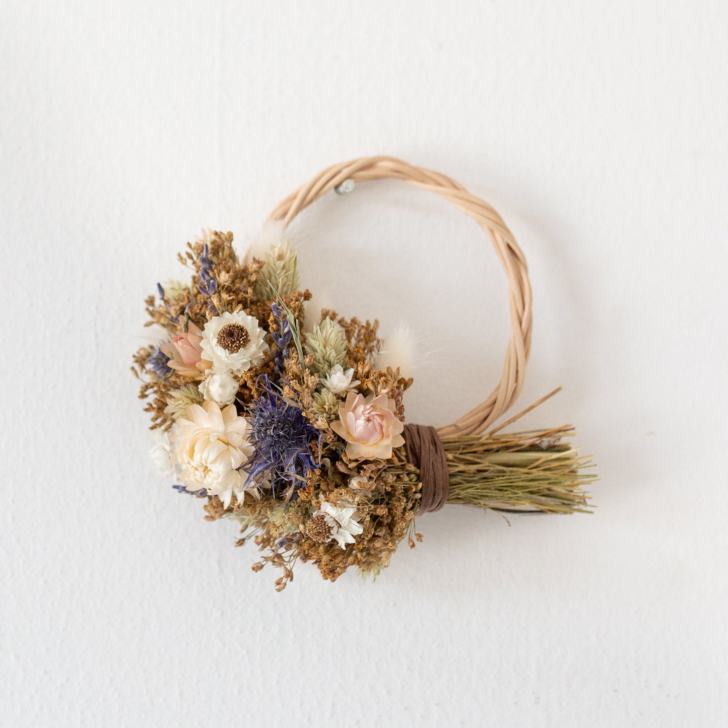 mini wreath wicker hoop half covered in white, blue and natural golden coloured dried flowers. The wreath is hung on a white wall