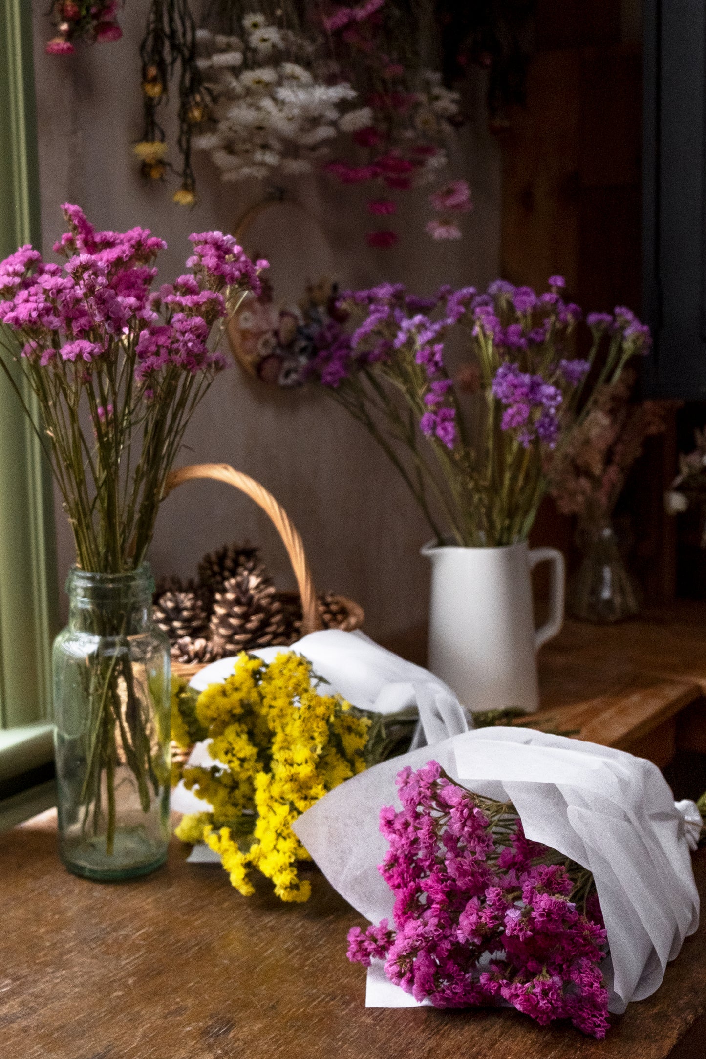 florist workspace with dried flowers hung in the background. dried pink and violet statice flowers in a vases and pink and yellow statice bunches laid on wooden table wrapped in white tissue paper