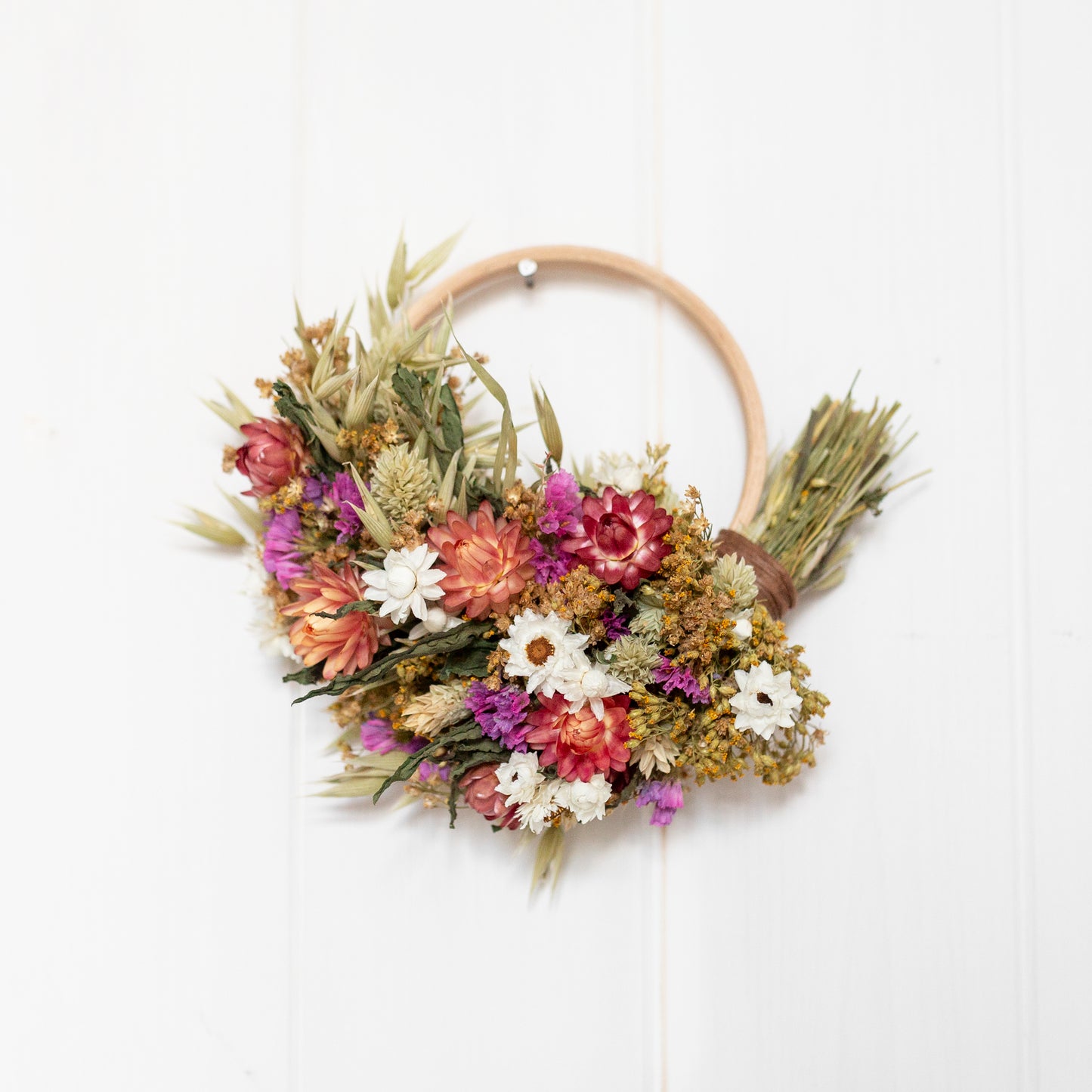 wooden hoop mini round wreath with green, white and pink dried flowers cover half the hoop. the wreath is hung on a white wood panelled wall