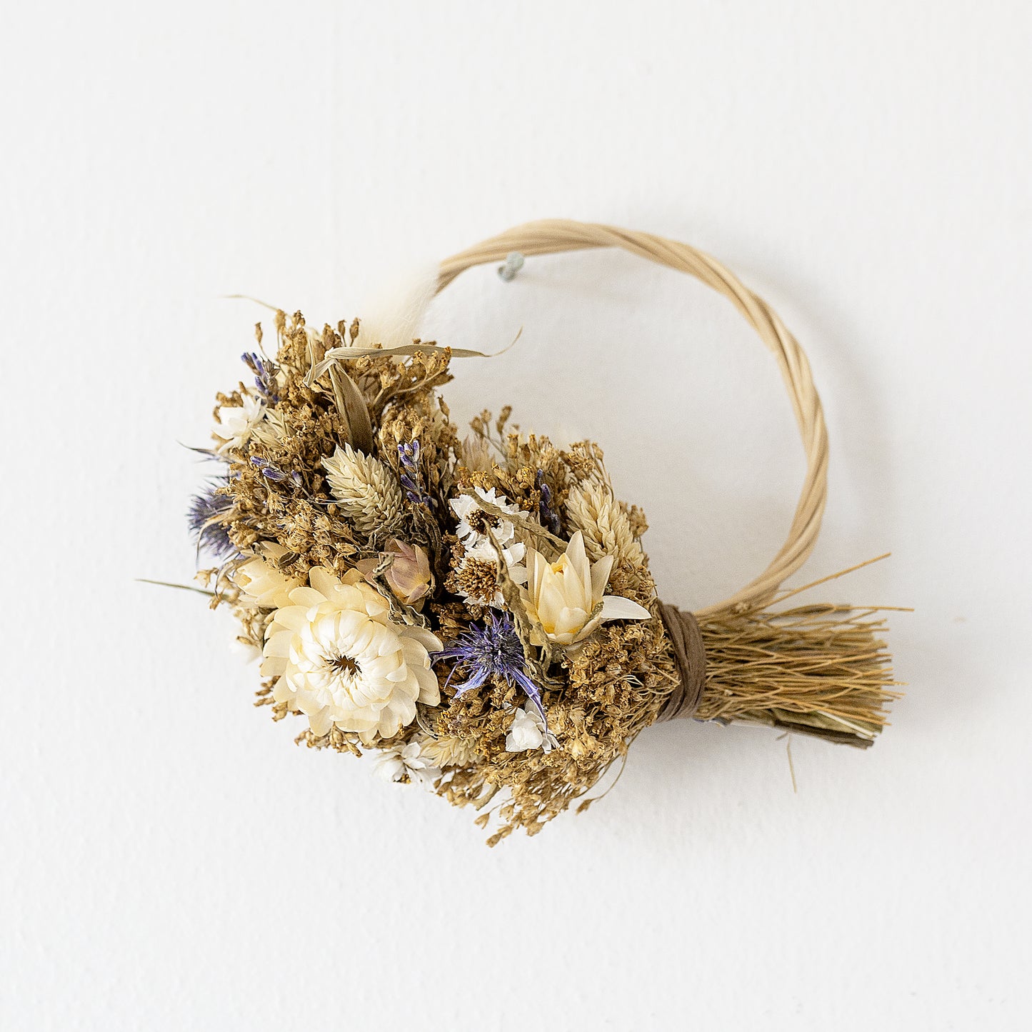 mini wicker wreath hung on a white wall, the wreath is half covered in dried flowers, white, blue, natural golden and pale green