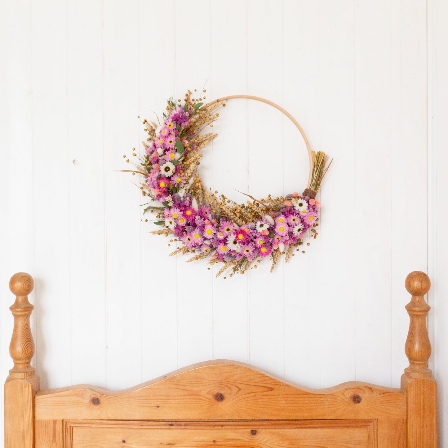 pink and lilac dried flower wreath hoop , wooden hoop half covered in flowers, hung up on a white panelled wall above a pine bed