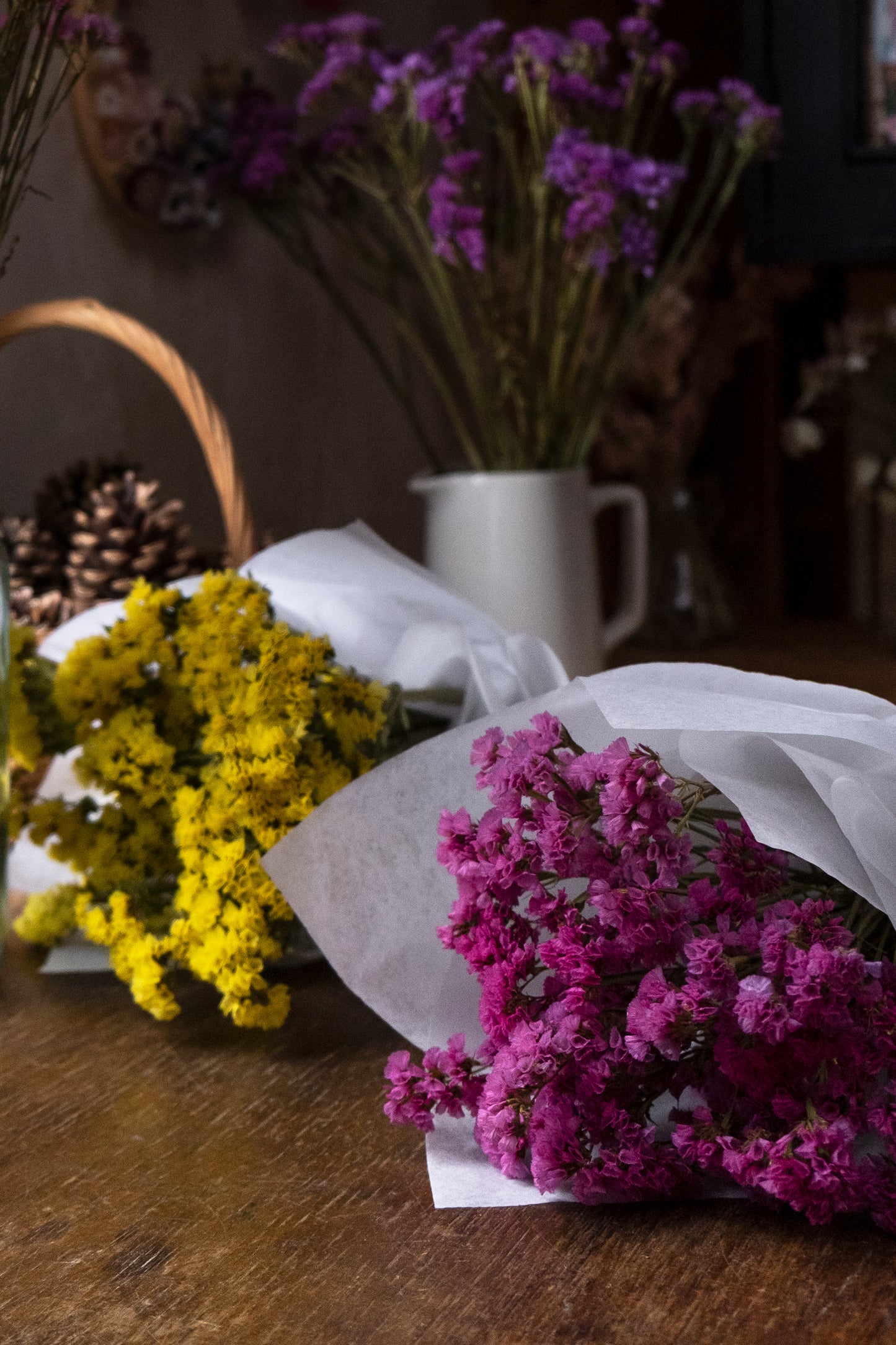 florist studio with violet statice in the background in a jug, yellow and pink statice in the front laid on a wooden table, the bunches are wrapped in paper