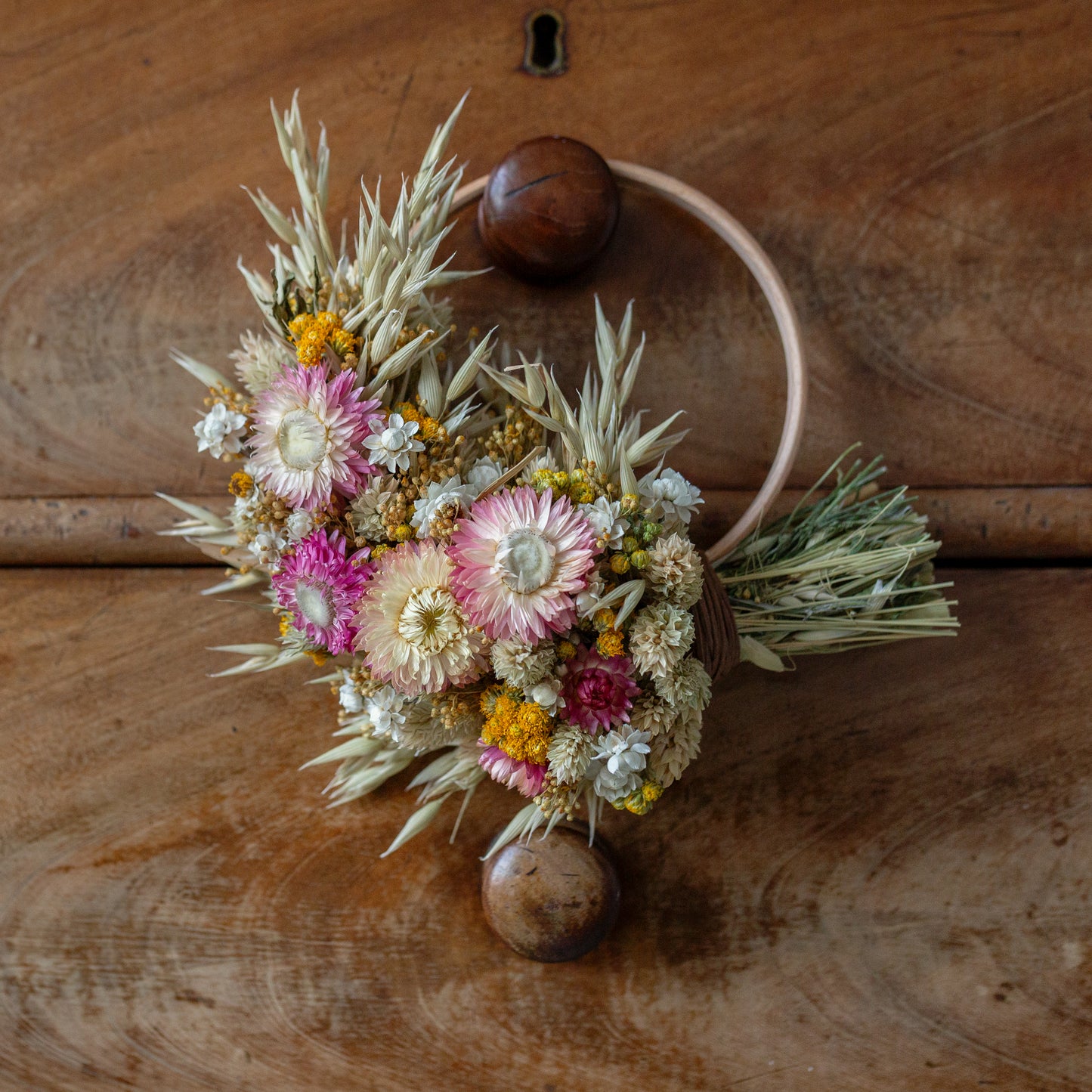 wooden wreath hoop half covered in dried flowers. pink strawflowers, yellow lona with oats and phalaris grass inbetween. the wreath is hung on a vintage brown drawer knob