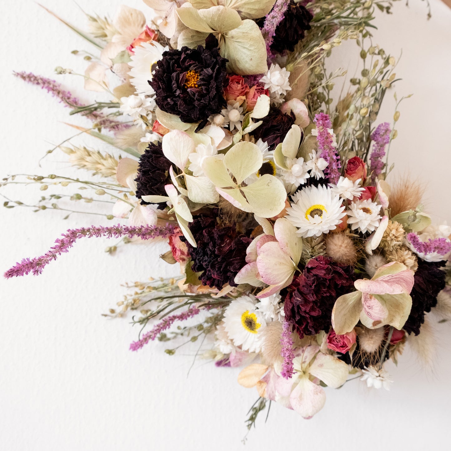 close up of side of a dried flower wreath on a white wall. flower and grass colours are green, white, dark red and purple