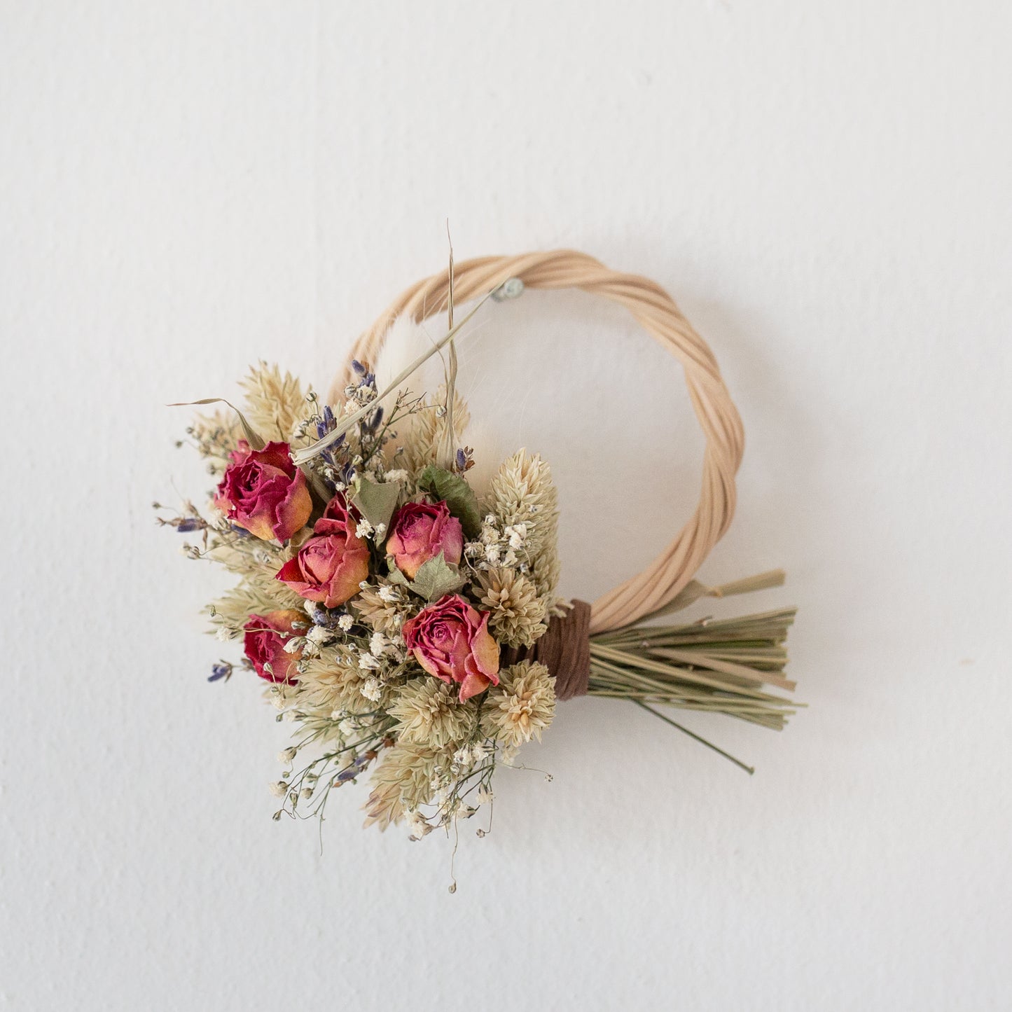 mini wicker wreath half covered in dried flowers, pink roses, lavender, Gypsophila. The wreath is hung on a white wall