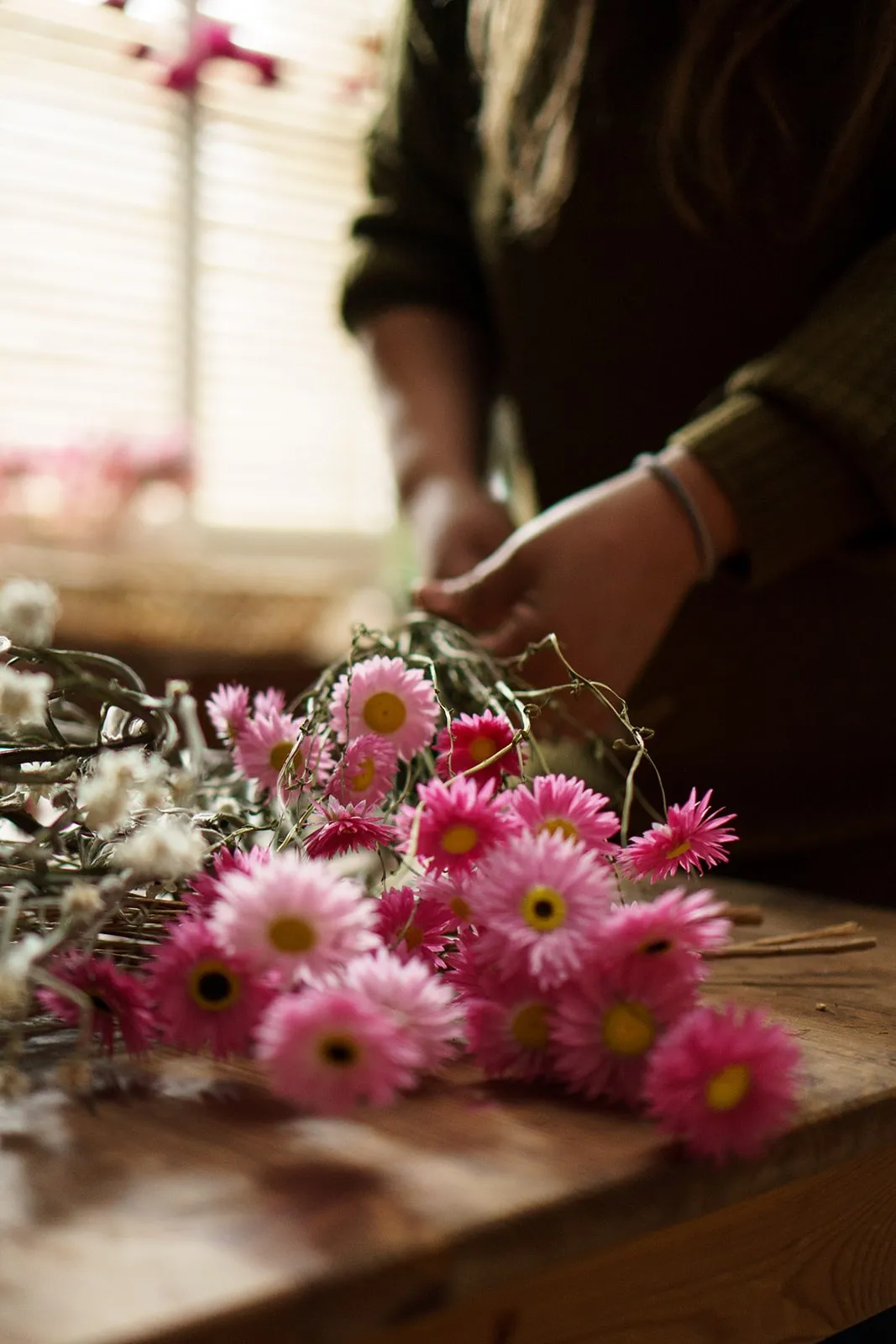 Hands arranging pink dried flowers on a wooden bench.