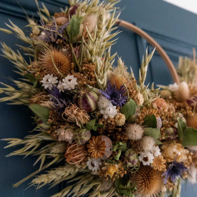 rustic dried flower wreath, neutral colours, greens, golden browns, with blue dotted around. the wreath is half covered in flowers and hung on a dark blue door