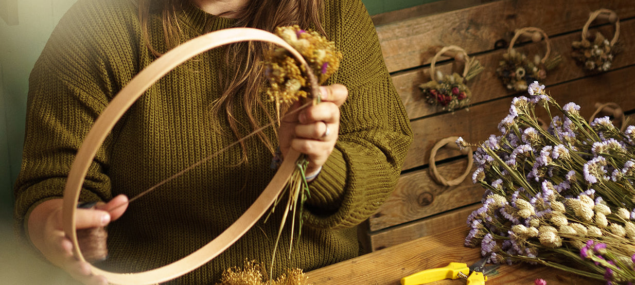 Jess at a book of petals holding a large wooden wreath hoop making a wreath with dried flowers against a rustic wooden background