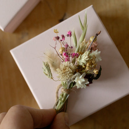 a dark pink, cream, and white dried flower mini posy tied together with natural twine in a hand on top of a pink gift box