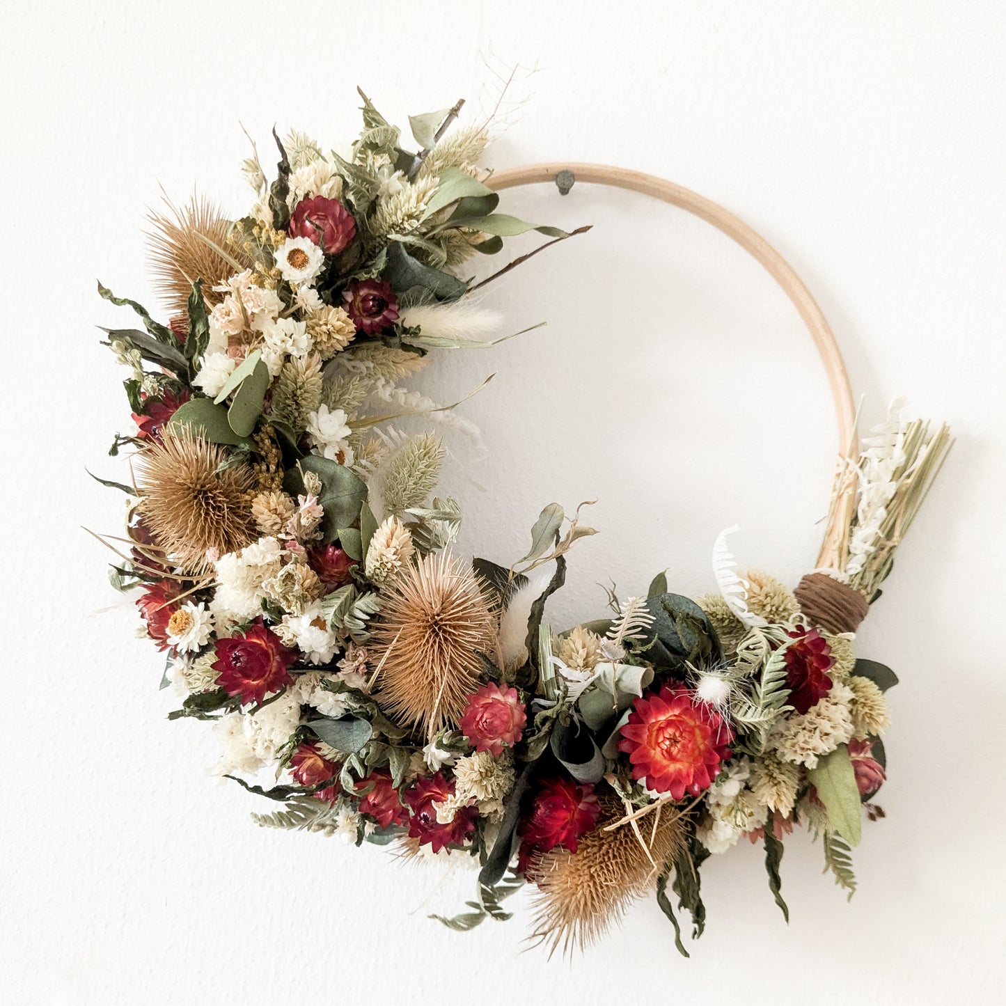 red, green, white and natural coloured dried flower wreath hoop. The hoop is hung on a nail on a white wall. The hoop is approx. 2/3 covered in flowers