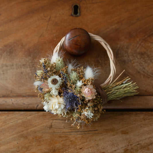 mini wreath wicker hoop half covered in dried flowers, white strawflowers, blue thistles, white bunny tails and golden bloom broom. the wreath is hung on a drawer knob