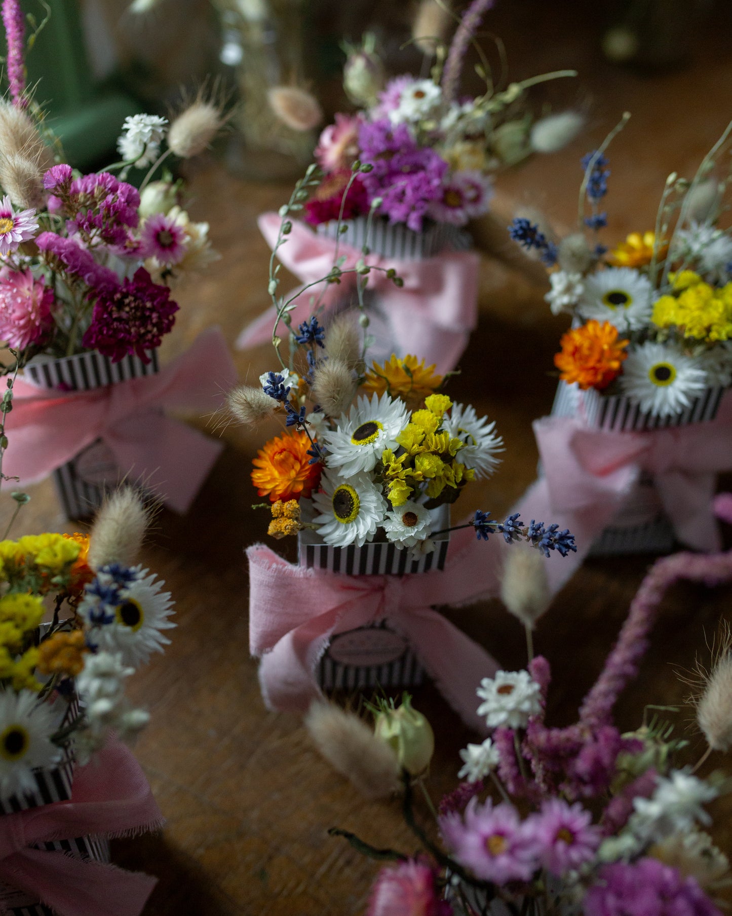dried flower posies in a stripy gift box tied with a pink cotton bow. 6 posies on a wooden table, 3 are pink flowers and 3 are mix white yellow and blue flowers