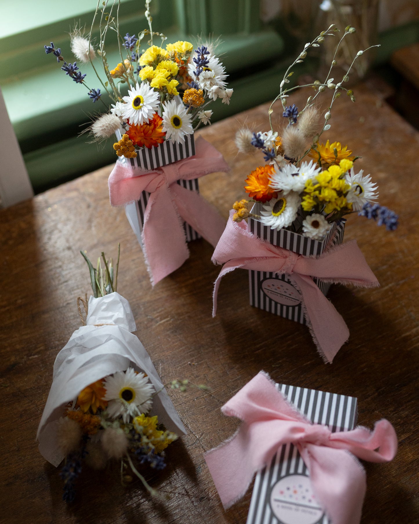 Small mixed coloured yellow white, orange and blue floral arrangements in striped boxes with pink bows on a wooden surface. 1 posy arrangement is wrapped in white tissue ready to post