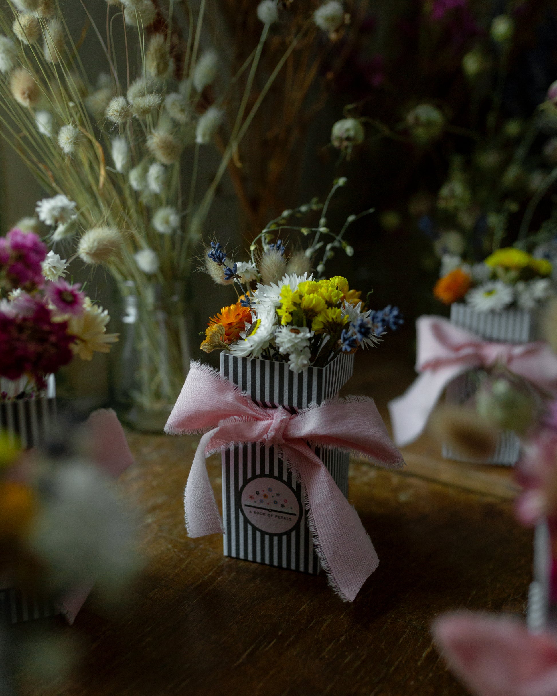 Small dried flower posy in a striped gift box with a pale pink bow, surrounded by other floral arrangements
