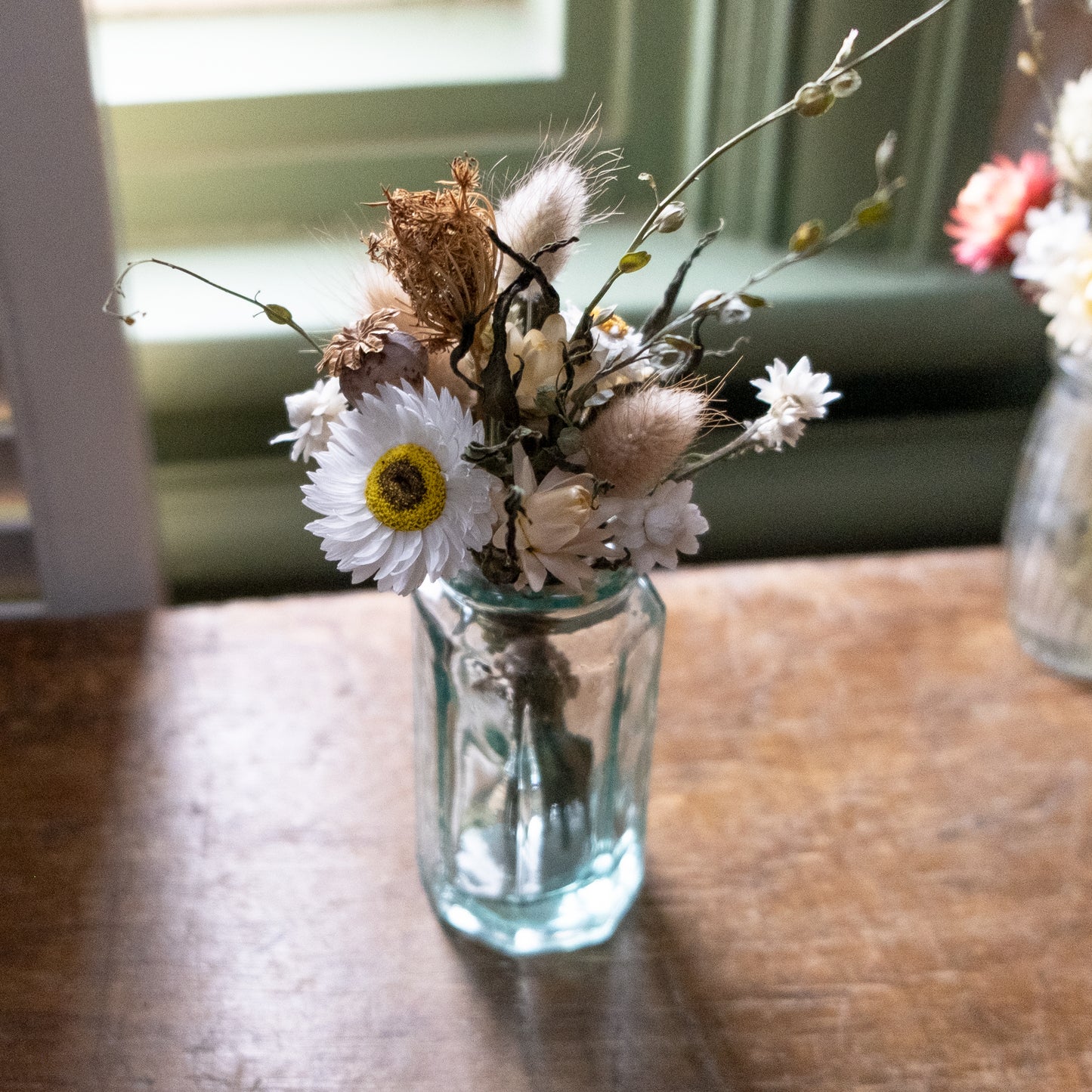 Clear glass bud vase with a white, green and light brown posy in on a wooden surface with a window in the background.