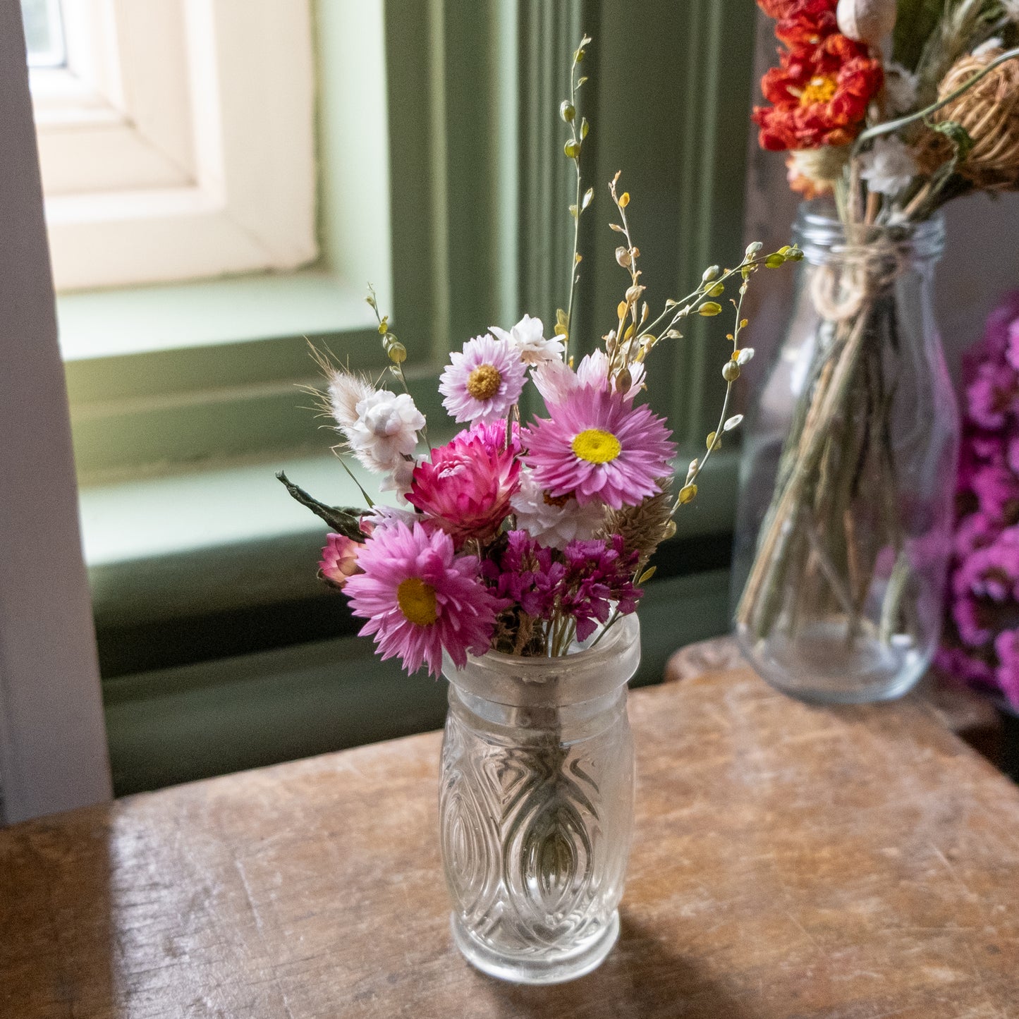 Clear glass  bud vase with pink flowers posy inside on a wooden surface near a green window.