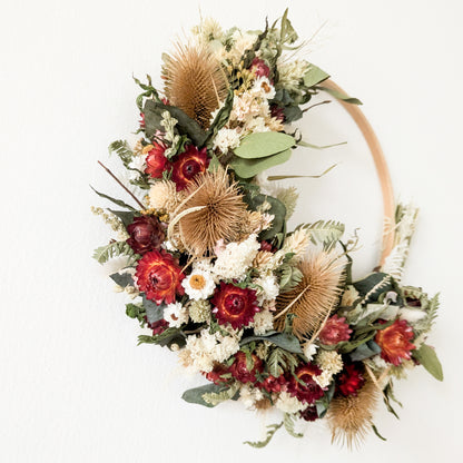 close up of a red, green, white and natural coloured dried flower wreath hoop. The hoop is hung on a nail on a white wall. 