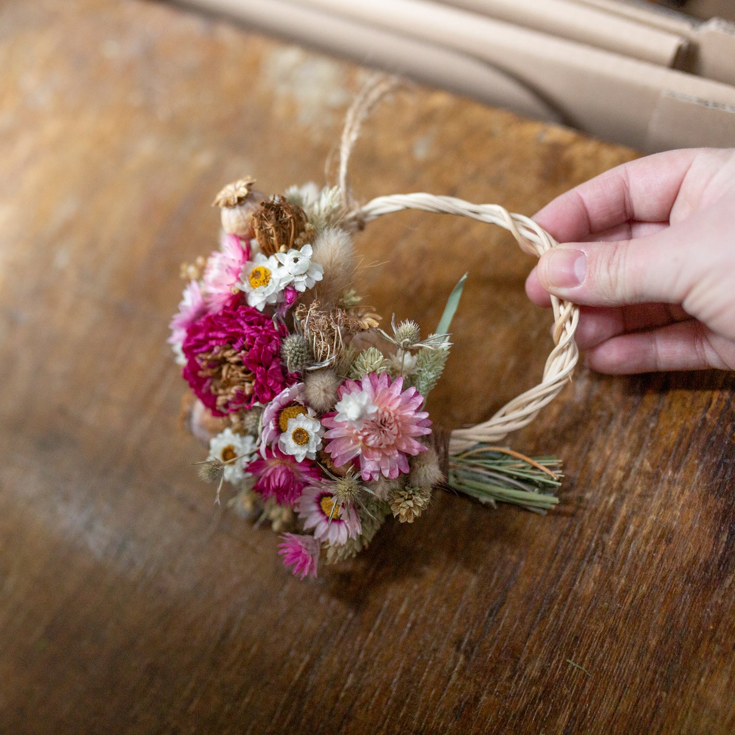 Hand holding a small dahlia and strawflower dried flower wreath of pink dried flowers on a wooden surface