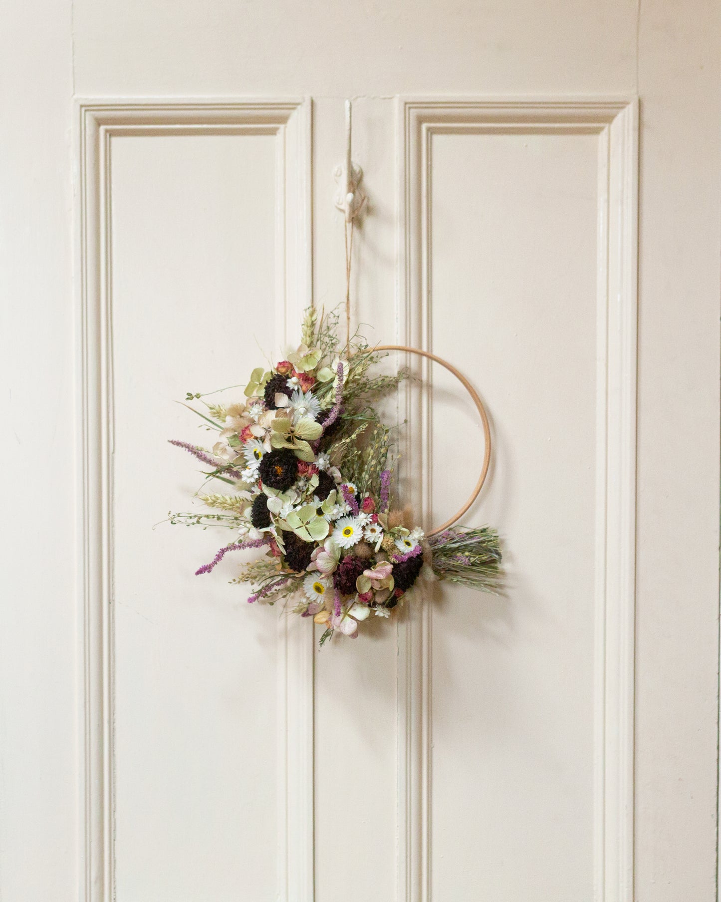 dahlias daisy hydrangea and grass half moon wreath. the wreath is made from dried flowers and a wooden hoop. The wreath is hung from twine on a hook on the back of a cream coloured door