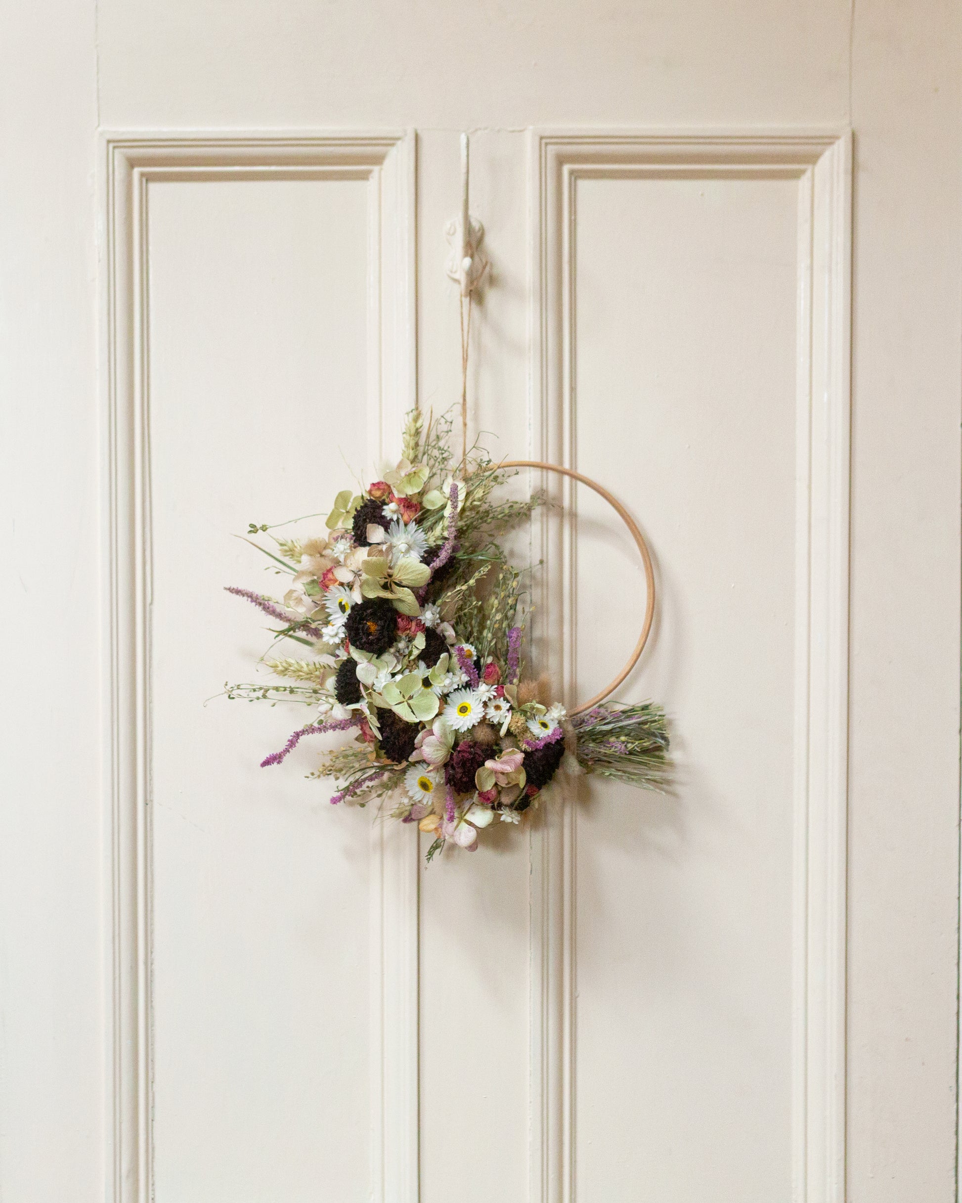dahlias daisy hydrangea and grass half moon wreath. the wreath is made from dried flowers and a wooden hoop. The wreath is hung from twine on a hook on the back of a cream coloured door