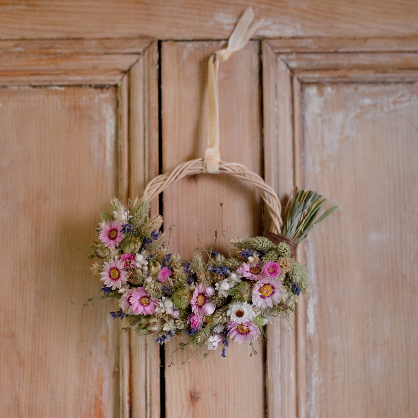 wicker hoop half covered in dried flowers hung on the back of a old wooden door. the wreath has pink and white daisies on and lavender 