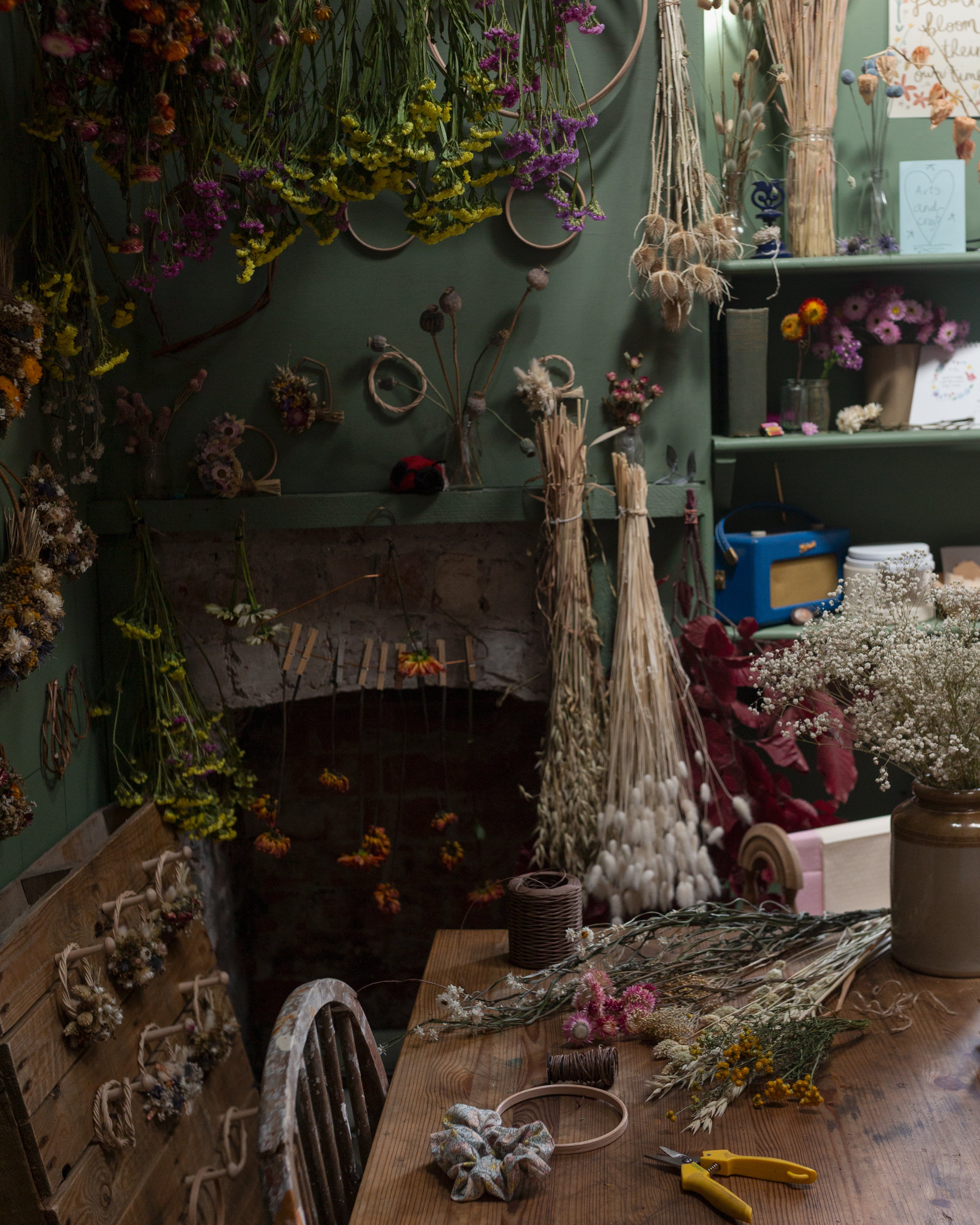 Floral workshop with dried flowers hanging from ceiling and in vases, dried flower wreaths on walls and wooden hooks. A green rustic setting.