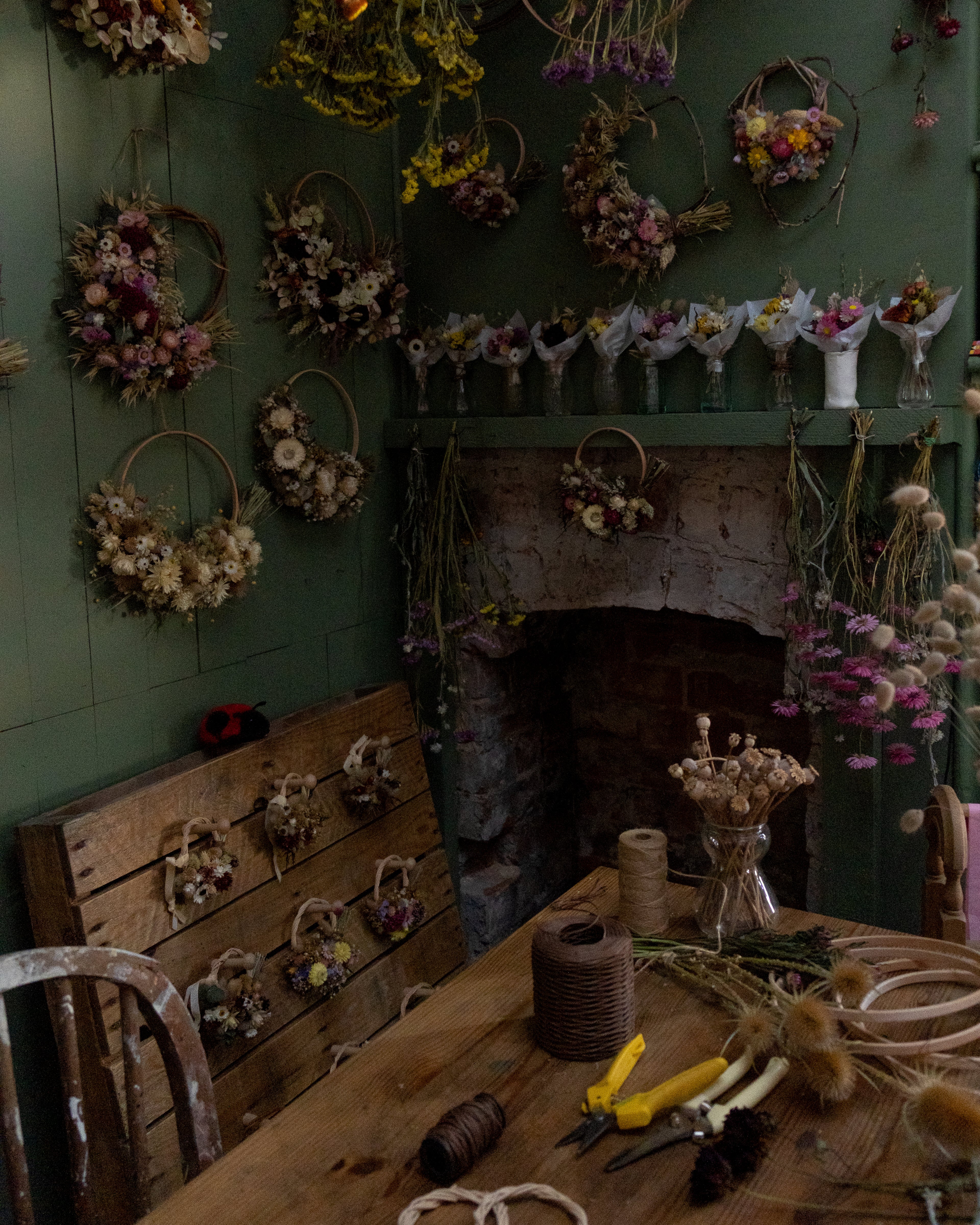 flower florist studio space with green walls and a open fireplace. the walls have dried flower wreath hanging and a mantle piece has small posies in jars. there is a wooden pallet with hooks with mini wreaths hanging. a work table with wire, hoops and flowers laid on ready to make