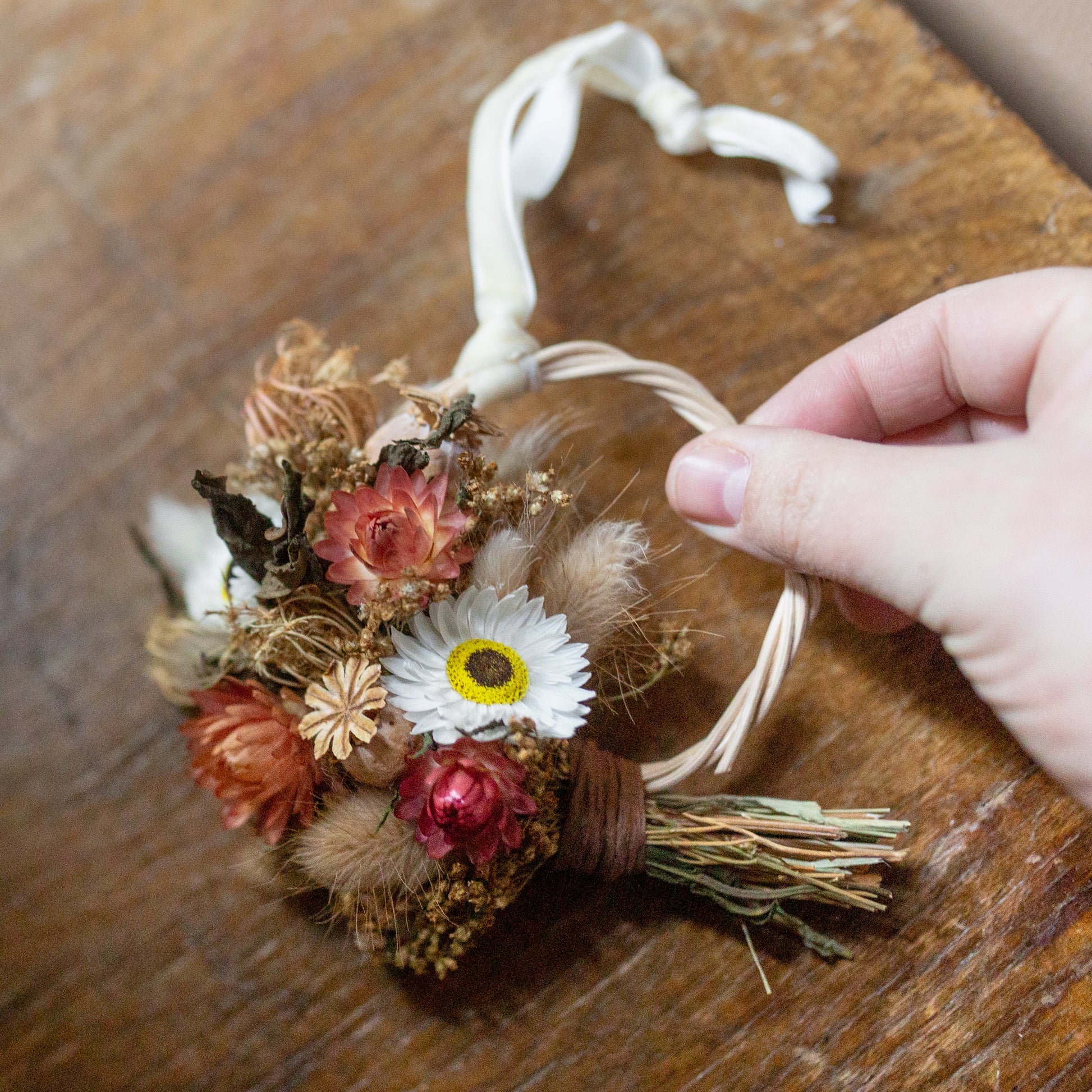 mini rattan wreath made from red white and golden dried flowers and grasses. the wreath has a creamy velvet ribbon attached for hanging and is held in a hand over a wooden table