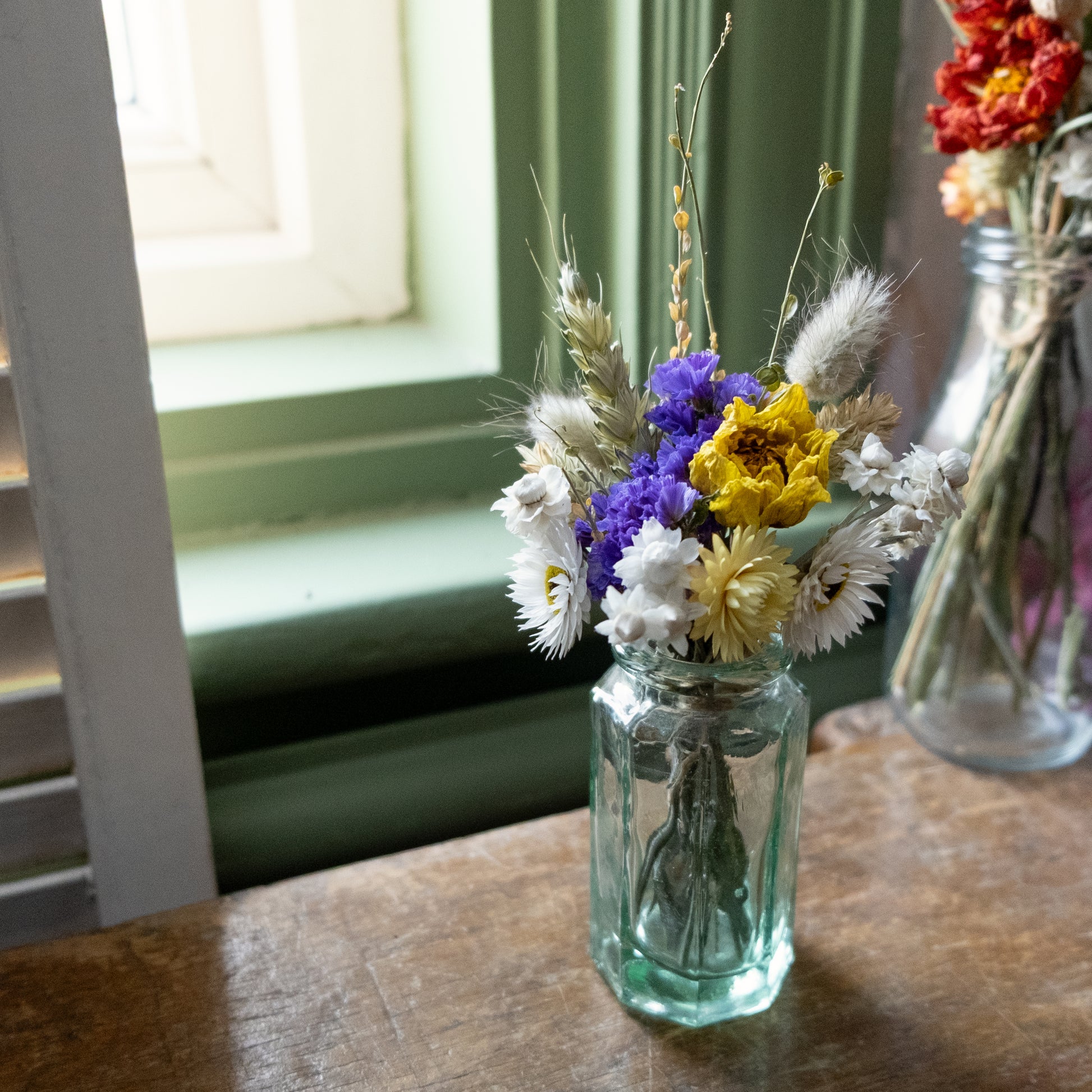 Clear glass bud vase with a white, yellow and blue flower posy in on a wooden surface with a window in the background.