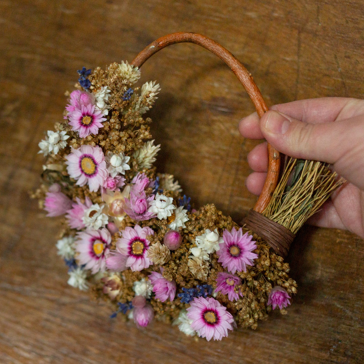small pink white and lavender dried flower wreath held in a hand over a wooden table