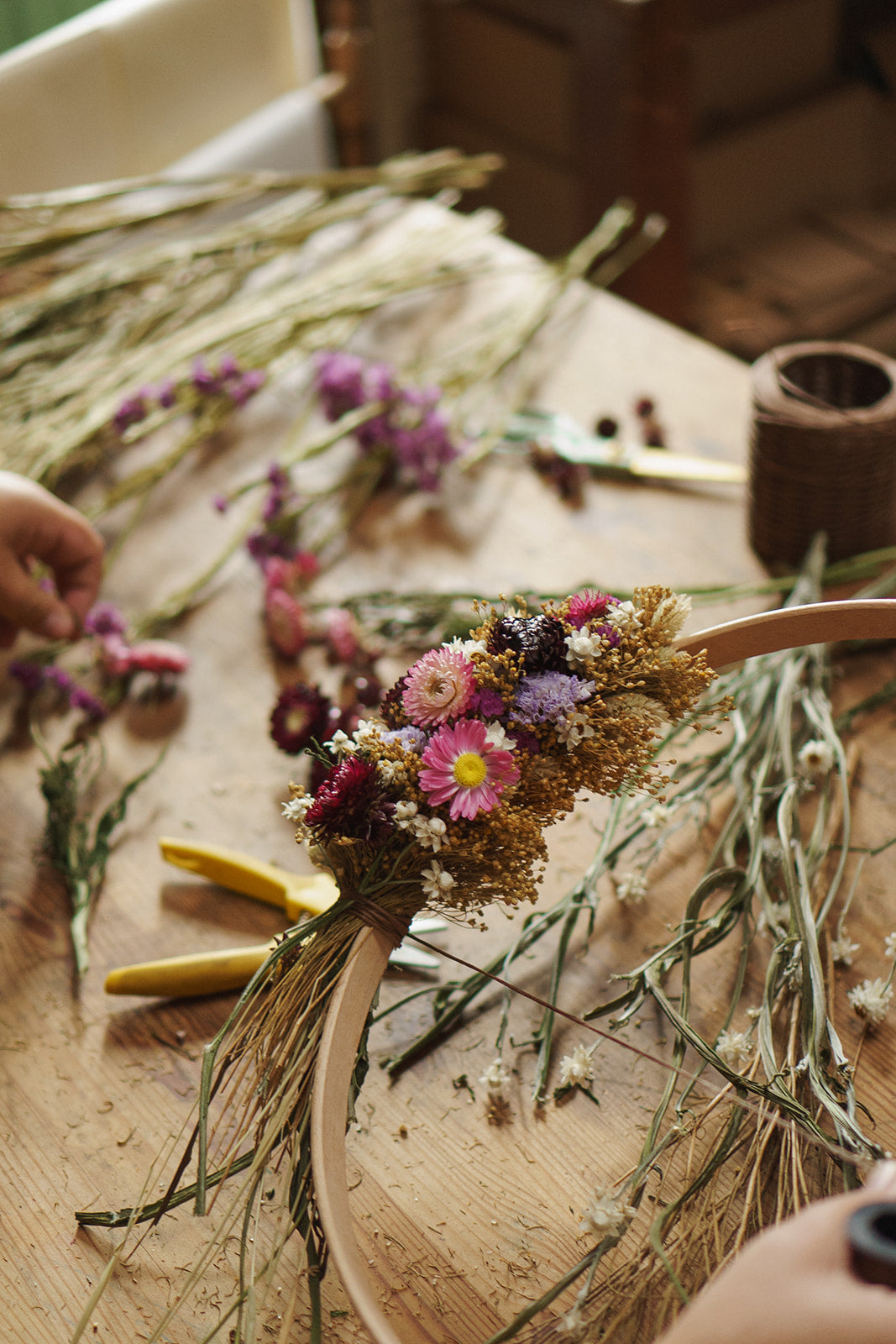 Dried flowers on a wooden surface with hands working on making a wreath wrapping flowers around a wooden hoop