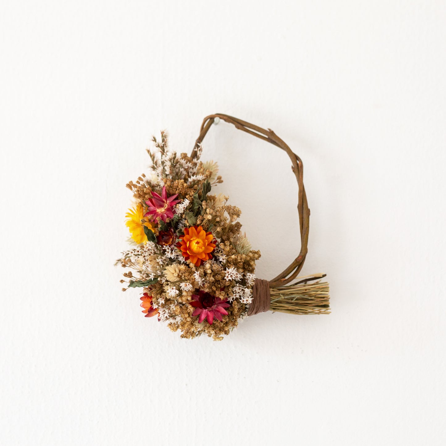 mini willow rustic style wreath, half covered in dried flowers, natural colours, red yellow orange and golden browns. the wreath is hung on a nail on a white wall