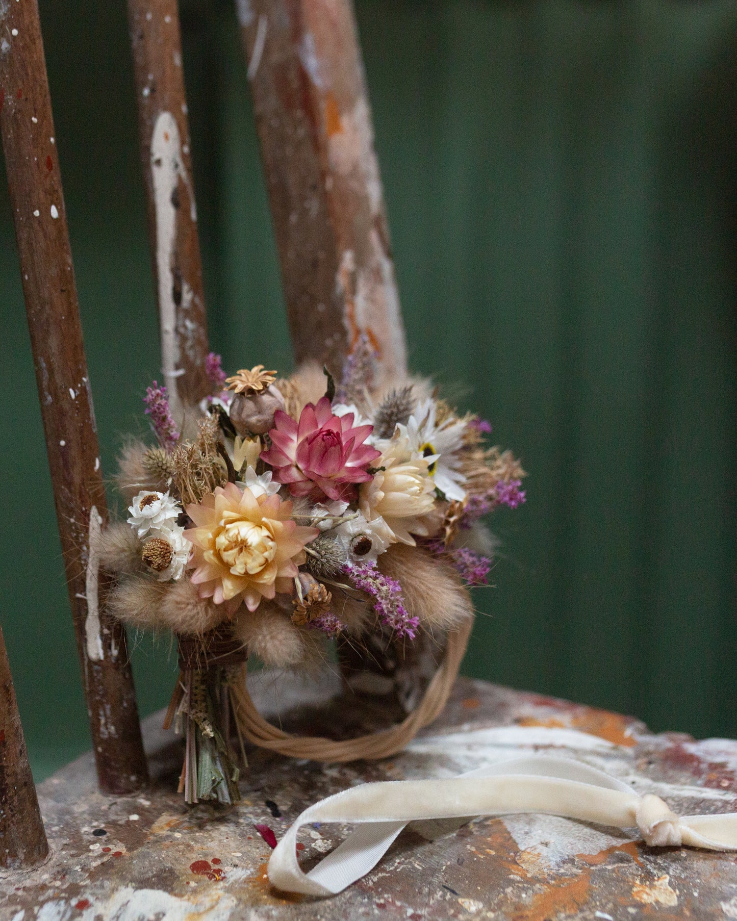 mini dried flower half moon wreath arrangement stood up leaning on the back of an old wooden chair with the flowers to the top and wicker base on the bottom, the chair has old paint splatters dried and is against a green background