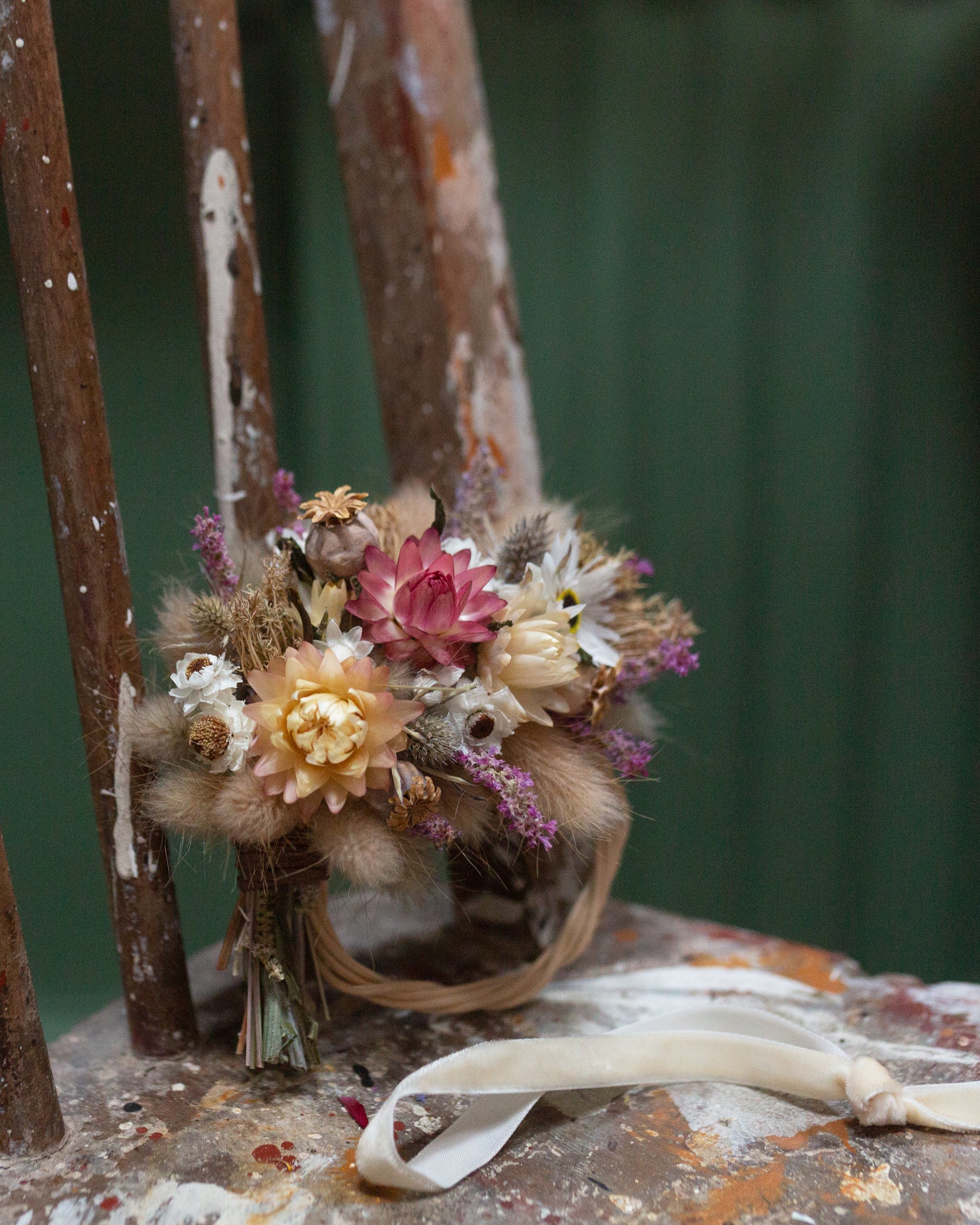 mini dried flower half moon wreath arrangement stood up leaning on the back of an old wooden chair with the flowers to the top and wicker base on the bottom, the chair has old paint splatters dried and is against a green background