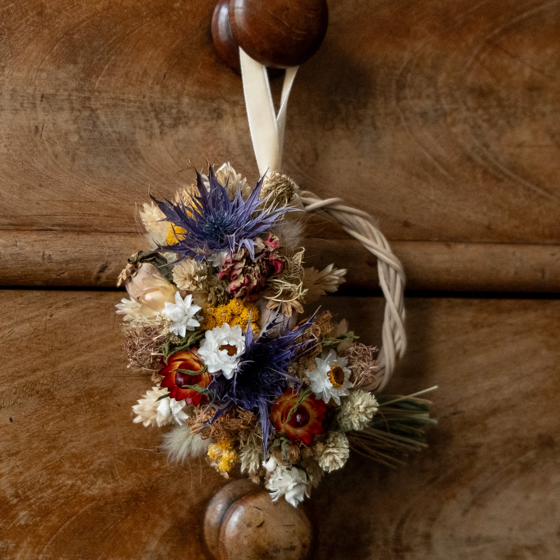 blue thistle, red zinnia and strawflower, yellow lona half moon mini wreath. the wreath base is make of rattan and is hung on a wooden drawer knob