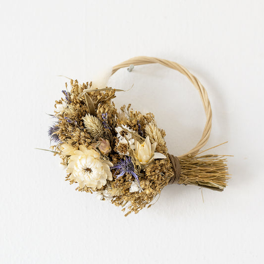 mini wicker wreath hung on a white wall, the wreath is half covered in dried flowers, white, blue, natural golden and pale green