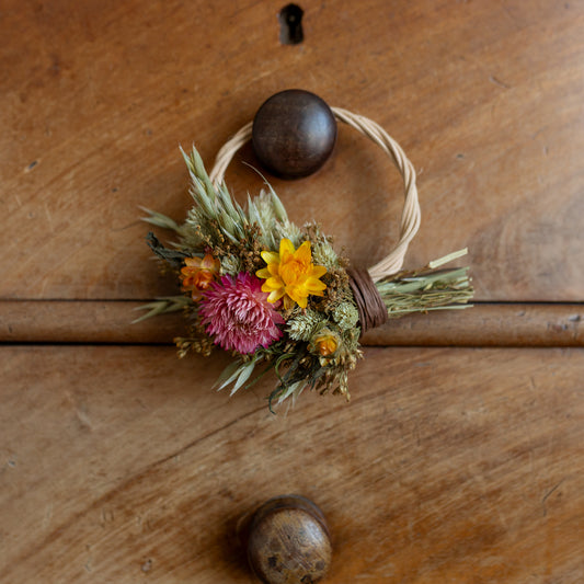 Small wicker wreath hoop, with green yellow and pink dried flowers.
the wreath is half covered with the flowers and is hung on a dresser drawer