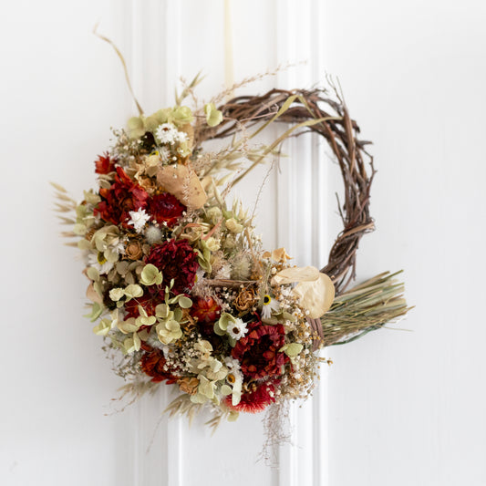 Decorative wreath with autumnal flowers and leaves on a white door. The wreath colours are red, cream and neutral colours