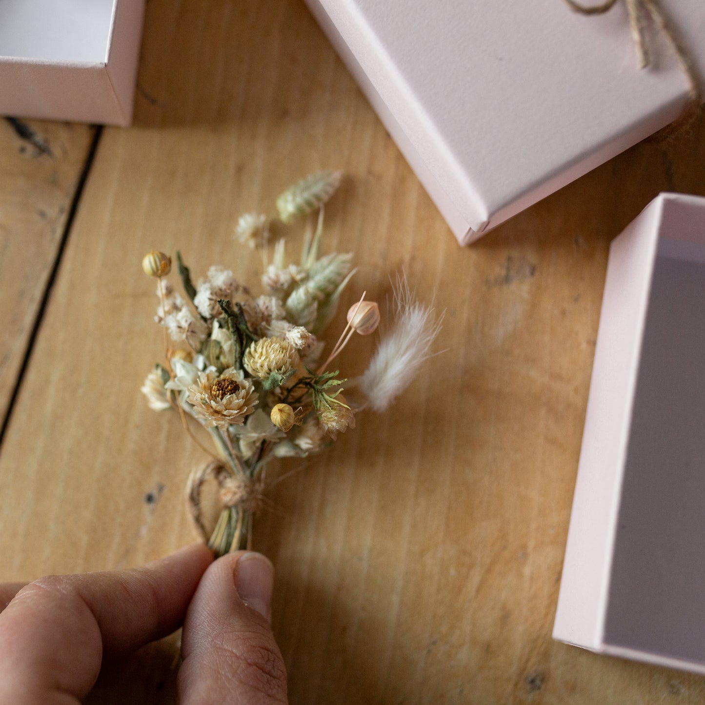 neutral coloured mini posy in fingers being place down on a table surrounded by pink gift boxes