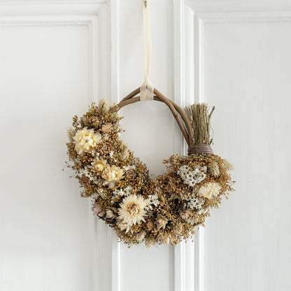 neutral dried flower wreath hung with a cream velvet ribbon on the back of a white door. the wreath is 3/4 covered , a homemade willow base, white strawflowers and ixodia daisies, natural golden broom bloom filler and creamy roses. 