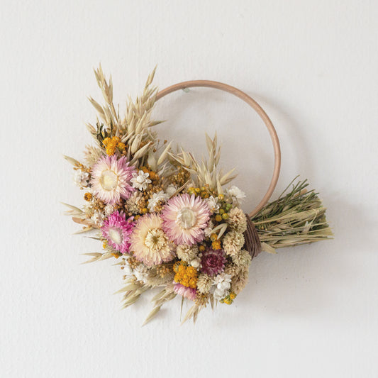 wooden wreath hoop half covered in dried flowers. pink strawflowers, yellow lona with oats and phalaris grass inbetween. the wreath is hung by a nail on a white wall