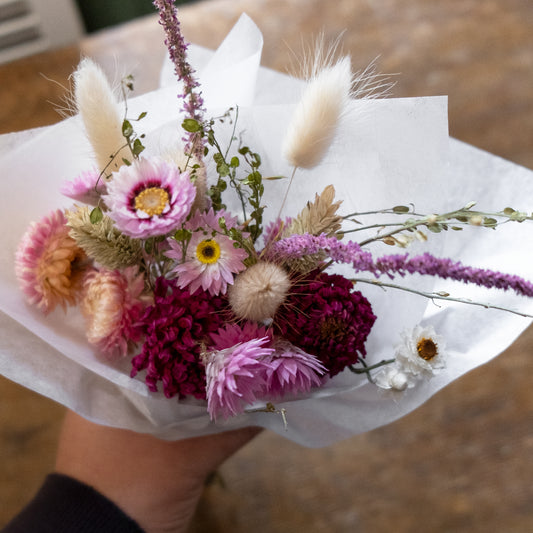 pink dried flower bouquet of flowers wrapped in white tissue held by a person with a blurred wooden background