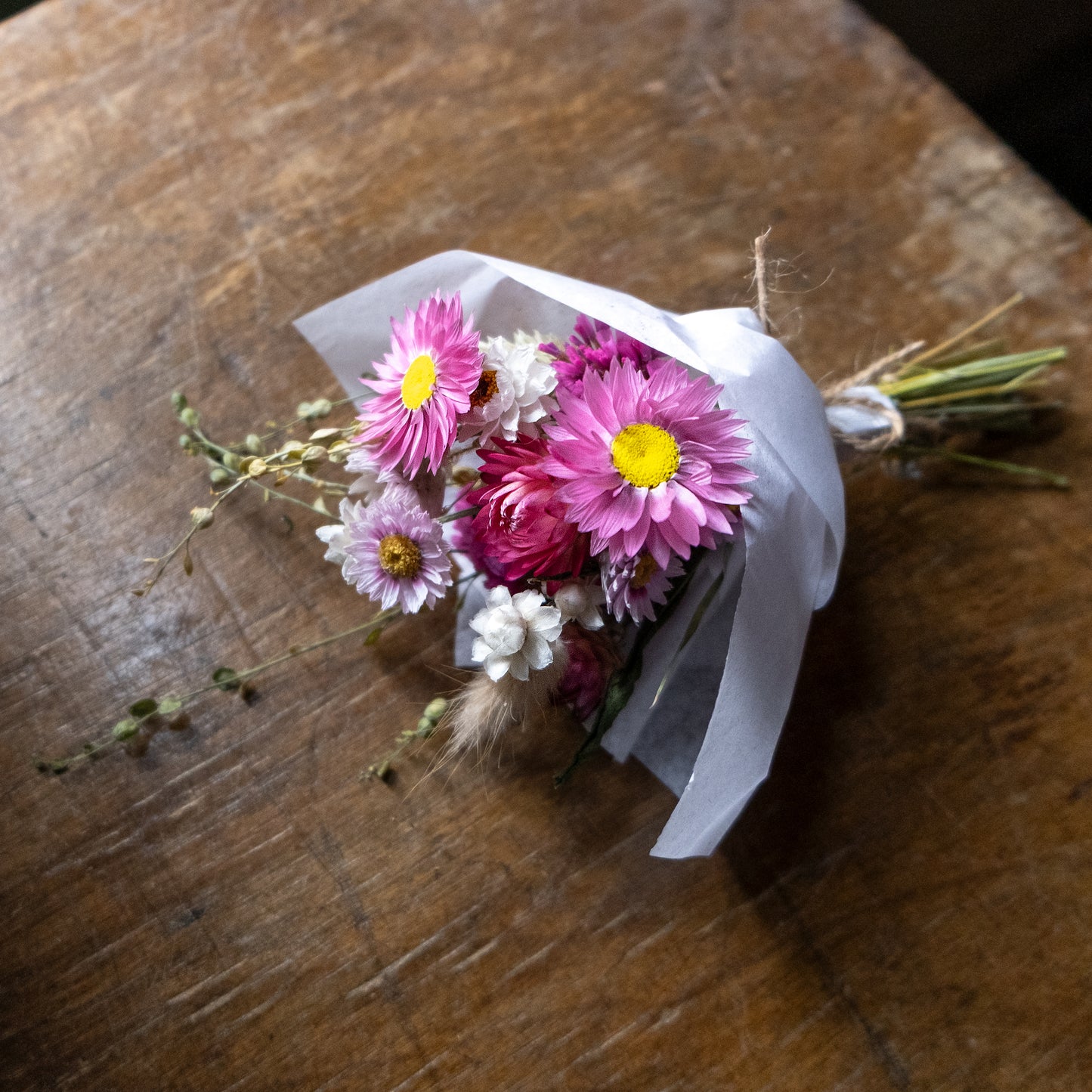 Small posy bouquet of dried pink and white flowers with grasses, wrapped in white tissue and tied with twine laid on a wooden surface