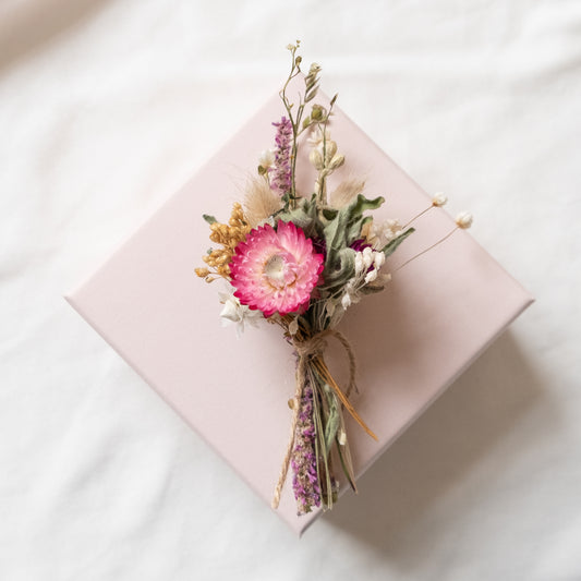 mini dried flower posy, made of pink and white flowers and grasses tied together with natural twine. the posy is laid on top of a pale pink gift box which is on a white background