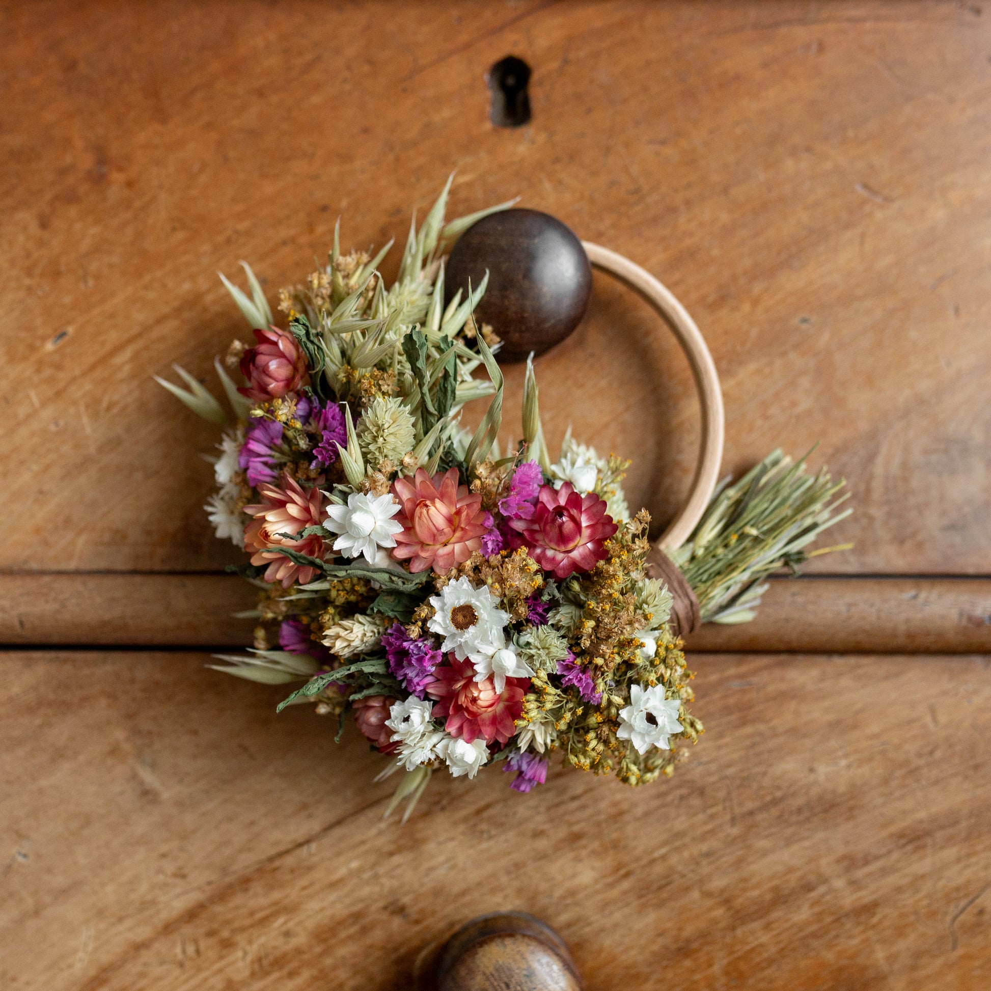 wooden hoop wreath with green, white and pink dried flowers cover half the hoop. the wreath is hung on a wooden drawer knob