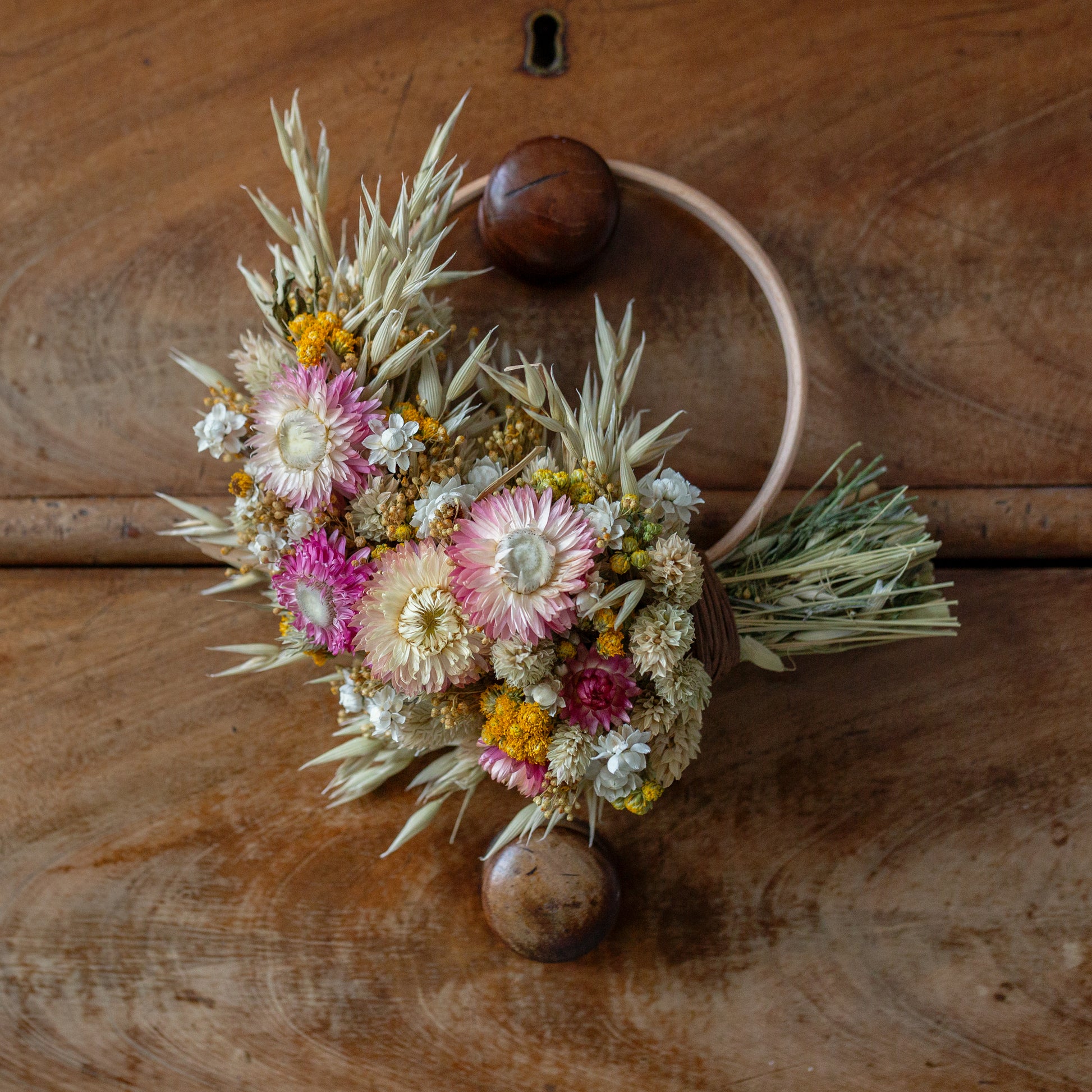 wooden wreath hoop half covered in dried flowers. pink strawflowers, yellow lona with oats and phalaris grass inbetween. the wreath is hung on a vintage brown drawer knob