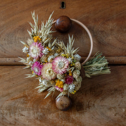 wooden wreath hoop half covered in dried flowers. pink strawflowers, yellow lona with oats and phalaris grass inbetween. the wreath is hung on a vintage brown drawer knob