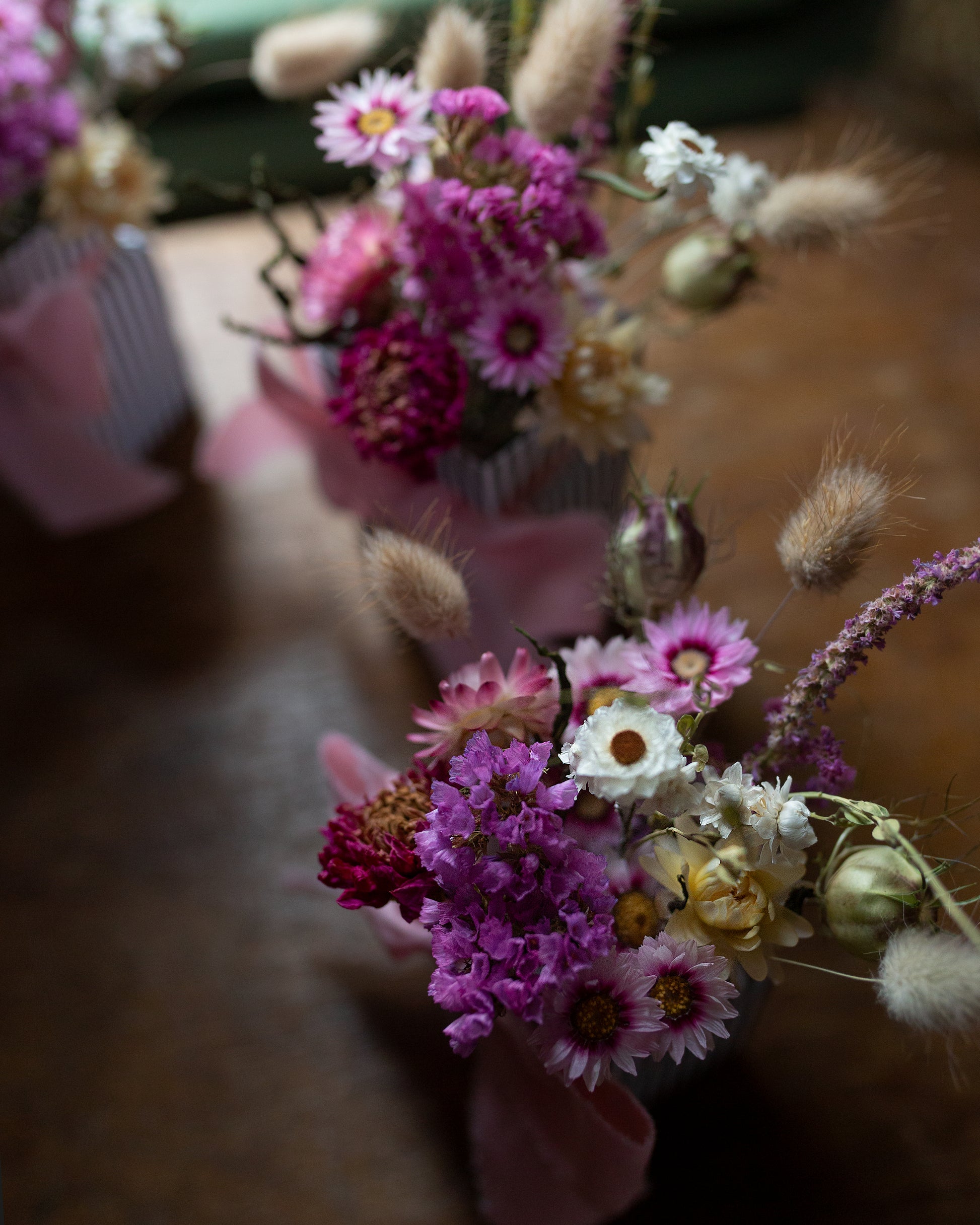 Bouquet of flowers with pink, white, and purpley pink colours on a blurred background bouquet in focus is made of dried flowers pink statice, pink and white strawflowers, dark pink dahlia bunny tails and grasses and white daisies nagella seed heads
