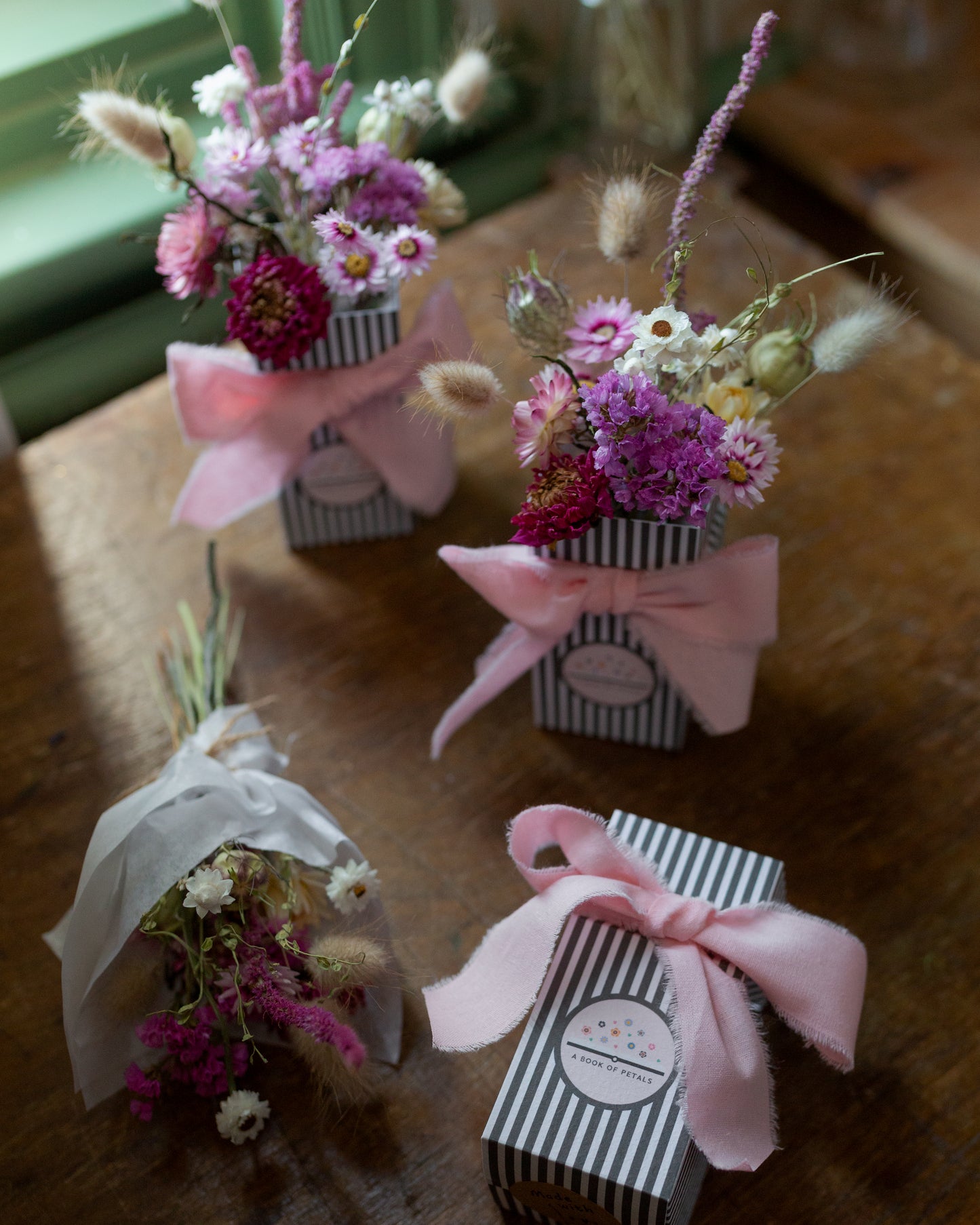 Small pink coloured floral arrangements in striped boxes with pink bows on a wooden surface. 1 posy arrangement is wrapped in white tissue ready to post